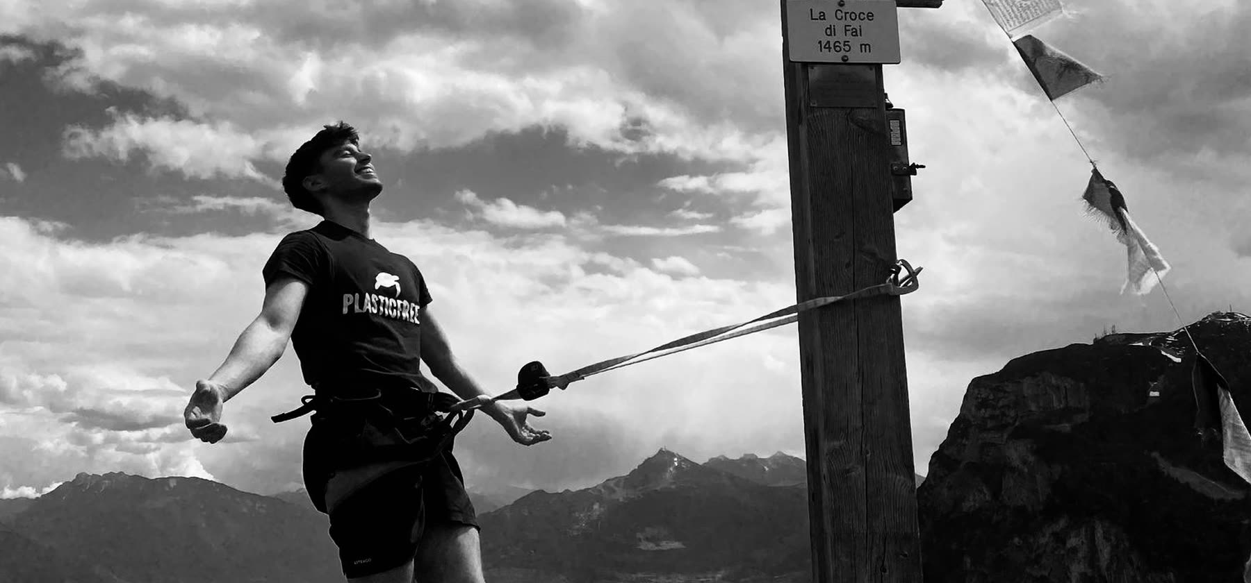 Markel leaning back on a rope at the summit cross of La Croce di Fai, mountains behind, in black and white